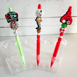 Może przedstawiać: Trzy ozdobione długopisy w przezroczystym plastikowym stojaku. Długopisy są czerwone i zielone z bożonarodzeniowymi ozdobami. Ozdoby obejmują czerwony kapelusz z napisem "Oh Fudge!" na nim, białą ceramiczną kulkę z napisem "A Christmas Story" na niej i zielono-czerwoną kulkę z napisem "You'll Shoot Your Eye Out Kid!" na niej.