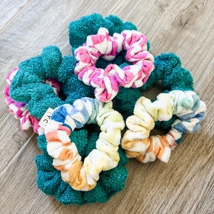 May include: A group of six colourful scrunchies made of soft, terry cloth fabric. The scrunchies are in a variety of colours and patterns, including pink and white stripes, rainbow stripes, and solid teal.