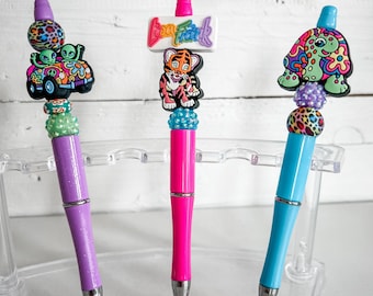 May include: Three colorful pens with decorative tops. The first pen has a purple body and a top with an alien in a car. The second pen has a pink body and a top with a tiger and the text "Boo Frank". The third pen has a blue body and a top with a turtle.