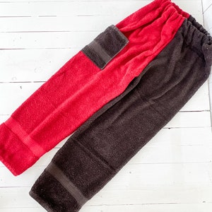 May include: Two terry cloth trousers, one red and one brown, are laid out on a white wooden surface. Each has a dark brown pocket and elastic waistband. The trousers appear to be made of a soft, absorbent material.
