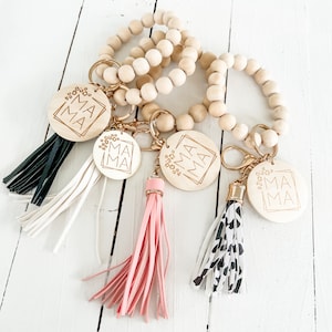 Pode incluir: Quatro pulseiras de contas de madeira com chaveiros dourados e borlas. Cada chaveiro tem um amuleto redondo de madeira com a palavra "MAMA" gravada em letras douradas. As borlas são pretas, brancas, cor-de-rosa e com padrão preto e branco.