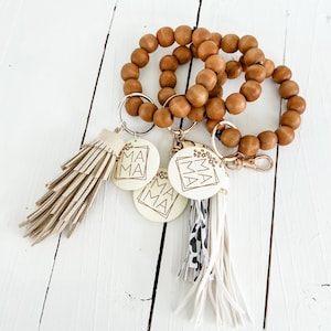 Pode incluir: Um chaveiro de contas de madeira com três etiquetas redondas de cor creme que dizem "Mama" em letras douradas. O chaveiro tem duas borlas, uma bege e uma preto e branco.
