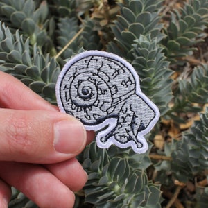 May include: Embroidered patch with a snail design. The patch has a grey body with black detailing and a white border. The snail's shell is intricately patterned. The patch is held against a background of green succulent plants.