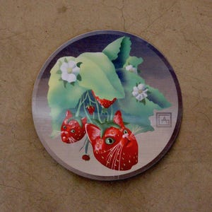 Puede incluir: Una pegatina circular con una ilustración surrealista de dos fresas rojas con forma de cabeza de gato, hojas verdes y flores blancas. Las fresas-gato tienen ojos verdes y bigotes blancos. Se observa un pequeño logotipo cuadrado con las letras AG.