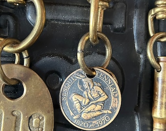 May include: A brass keychain featuring a round commemorative coin stamped with the text SESQUICENTENNIAL 1820-1970 and an image of a kneeling figure. The coin is attached to a brass swivel hook alongside a brass tag with the number 119 and a bullet casing.