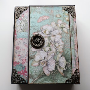 May include: A decorative journal with a floral design featuring white orchids and a butterfly on a teal background. The journal has a brown leather cover with brass corners and a brass knob. The text "My Book" and "Fleurs" is visible on the cover.