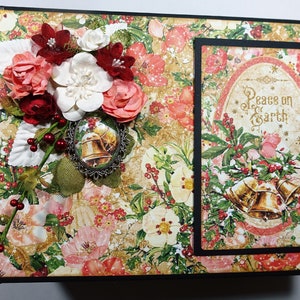 以下が含まれることがあります： 花柄のデザインと「Peace on Earth」という文字が書かれた金色と白の楕円形が特徴のクリスマスをテーマにしたスクラップブックの表紙。表紙は赤い実、緑の葉、金色のベルで飾られています。