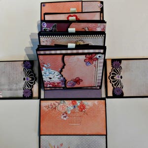 May include: A multi-layered scrapbook with a floral theme. The scrapbook is decorated with purple flowers, a clock face, and a bird. The scrapbook is closed and has a grey and white patterned cover.