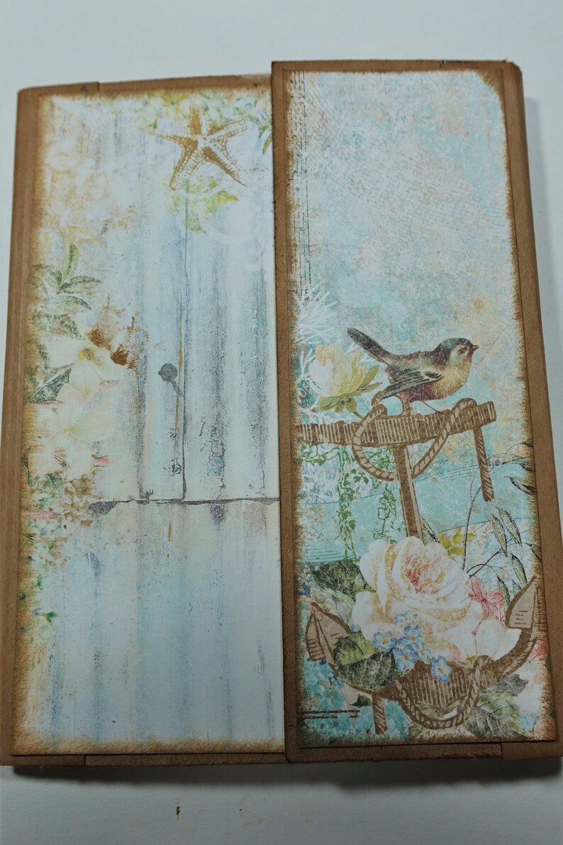 以下が含まれることがあります： 青と白の背景に、錨に止まっている鳥、バラ、ヒトデが描かれたスクラップブッキングペーパー。紙は、茶色の縁取りで、使い古したような風合いになっている。