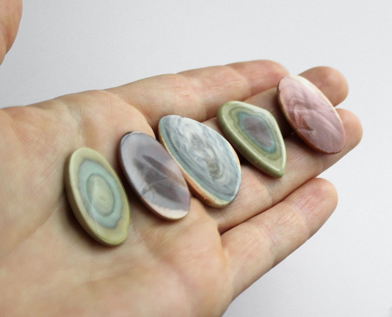 May include: Five polished, oval-shaped stones with a variety of colors and patterns, including green, brown, and pink. The stones are arranged in a row on a hand.