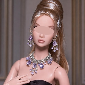 Peut inclure: Une poupée aux cheveux blonds coiffés en chignon, portant un collier argenté et des boucles d'oreilles assorties. Les bijoux présentent des cristaux violets clairs en forme de poire, entourés de petites pierres claires sur monture.