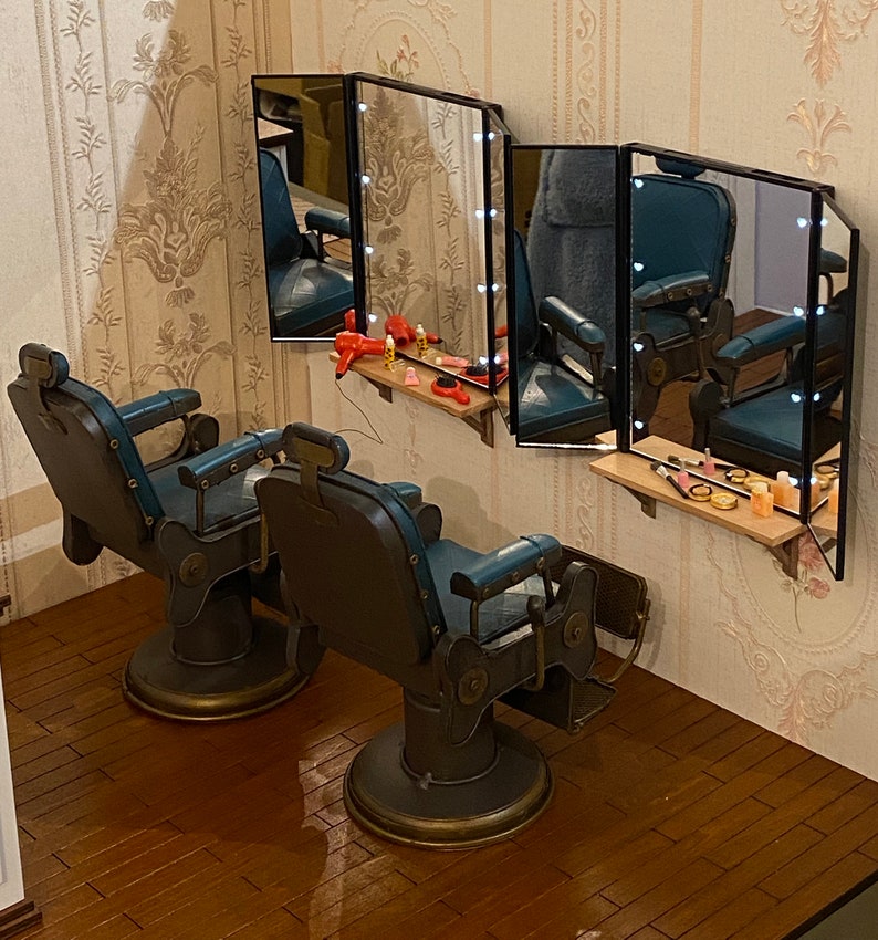 K&ouml;nnte beinhalten: Zwei Vintage-Friseurst&uuml;hle mit blauer Polsterung und Messingakzenten. Die St&uuml;hle stehen vor einem Schminkspiegel mit mehreren Lichtern. Der Spiegel ist von einem Holzrahmen umgeben.