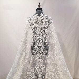 Puede incluir: Tela de encaje blanco con un intrincado dise&ntilde;o floral y de volutas. La tela est&aacute; drapeada sobre un maniqu&iacute;, mostrando el patr&oacute;n detallado. El encaje es adecuado para vestidos de novia o de gala.