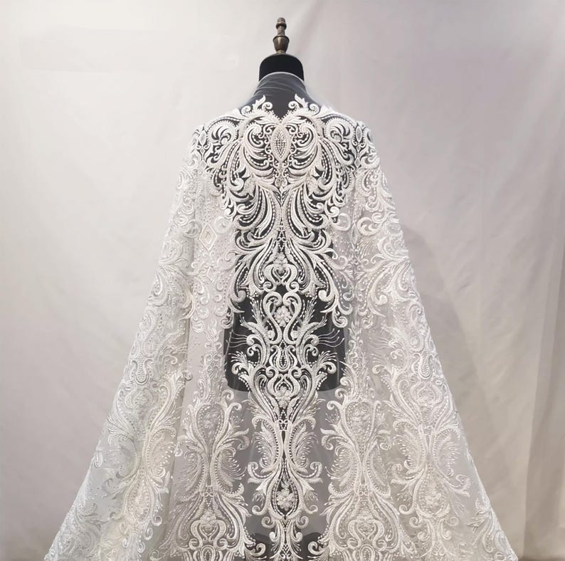 Puede incluir: Tela de encaje blanco con un intrincado dise&ntilde;o floral y de volutas. La tela est&aacute; drapeada sobre un maniqu&iacute;, mostrando el patr&oacute;n detallado. El encaje es adecuado para vestidos de novia o de gala.