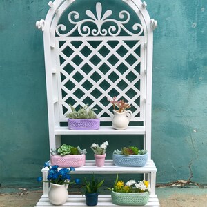 Puede incluir: Un soporte para plantas de madera blanca con tres estantes. El soporte tiene un dise&ntilde;o de enrejado en el estante superior y est&aacute; decorado con un dise&ntilde;o floral. Los estantes est&aacute;n llenos de plantas en macetas y una jarra blanca.