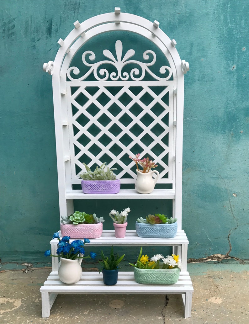 Puede incluir: Un soporte para plantas de madera blanca con tres estantes. El soporte tiene un dise&ntilde;o de enrejado en el estante superior y est&aacute; decorado con un dise&ntilde;o floral. Los estantes est&aacute;n llenos de plantas en macetas y una jarra blanca.