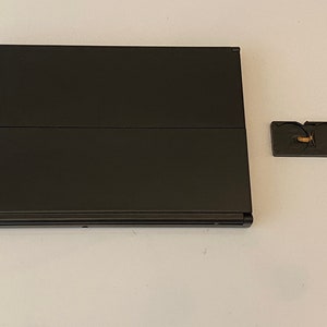 K&ouml;nnte beinhalten: Ein schwarzer Laptop mit geschlossenem Deckel. Der Laptop steht auf einer wei&szlig;en Oberfl&auml;che. Ein kleines schwarzes Zubeh&ouml;rteil mit goldenem Detail befindet sich rechts neben dem Laptop.