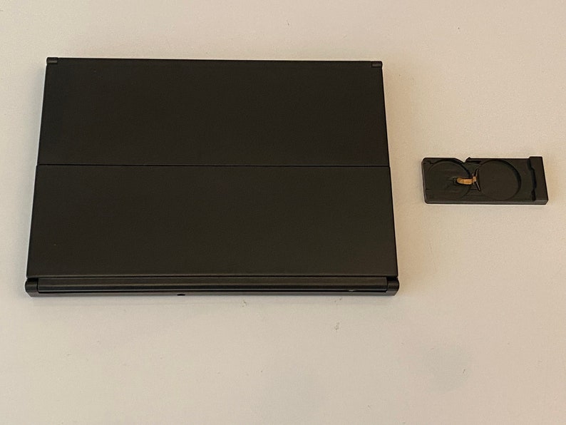 K&ouml;nnte beinhalten: Ein schwarzer Laptop mit geschlossenem Deckel. Der Laptop steht auf einer wei&szlig;en Oberfl&auml;che. Ein kleines schwarzes Zubeh&ouml;rteil mit goldenem Detail befindet sich rechts neben dem Laptop.