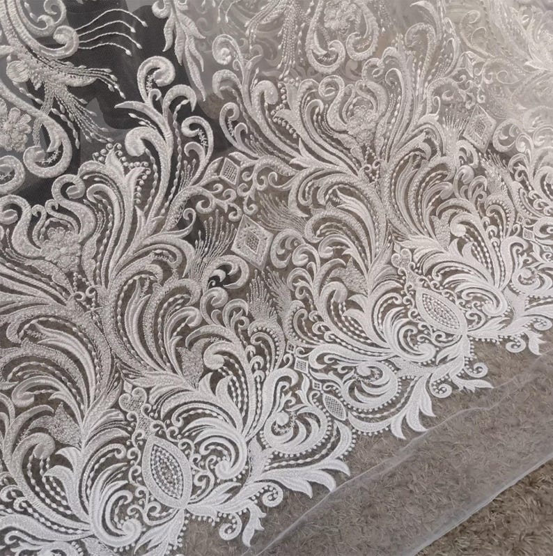 Puede incluir: Primer plano de tela de encaje blanco con un intrincado dise&ntilde;o floral y de volutas. El delicado patr&oacute;n presenta bordados detallados con una variedad de formas, incluyendo hojas, espirales y acentos en forma de diamante. El encaje tiene una calidad transparente y aireada.