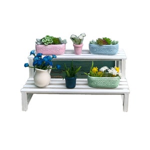 Puede incluir: Un soporte para plantas de madera blanca de dos niveles con cinco plantas en macetas. Las plantas incluyen suculentas, una planta verde y flores azules y amarillas. Las macetas son rosas, azules, verdes y blancas.