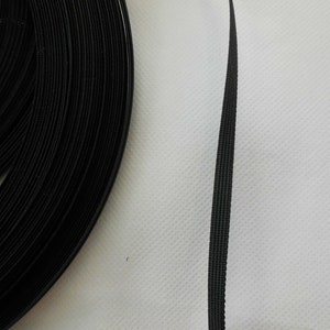 Puede incluir: Un rollo de cinta de gancho y bucle negro, tambi&eacute;n conocida como Velcro, se muestra sobre una superficie blanca. La cinta est&aacute; enrollada y una sola tira se extiende para mostrar la textura del material.