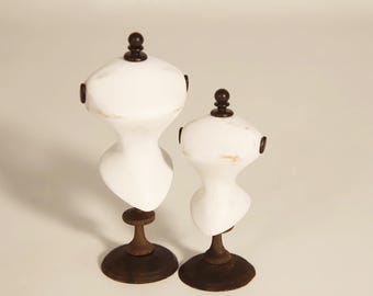 May include: Two white, vintage-style dressmaker forms with dark brown bases and finials. The forms are in different sizes, showcasing a classic design. The forms are ideal for displaying or creating clothing.