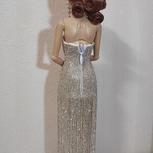 May include: A doll in a strapless, silver beaded gown with a long fringe. The doll has auburn hair in an updo. The dress has a back zipper. The doll stands on a black base.
