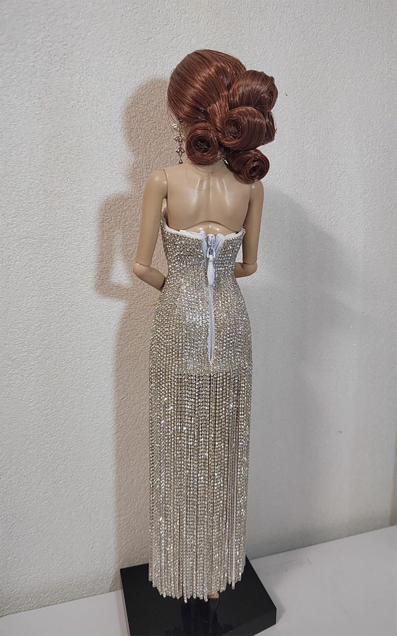 May include: A doll in a strapless, silver beaded gown with a long fringe. The doll has auburn hair in an updo. The dress has a back zipper. The doll stands on a black base.