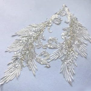 May include: Two ornate, symmetrical appliques with a feather-like design. The off-white fabric is embellished with silver sequins, creating a sparkling effect. The appliques are likely for embellishing garments or crafts.