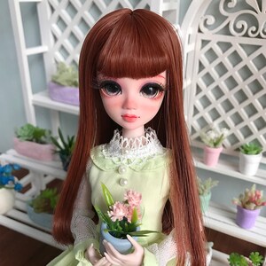 Puede incluir: Una mu&ntilde;eca con el pelo largo, liso y casta&ntilde;o, que lleva un vestido verde claro con un cuello de encaje blanco y sostiene una maceta peque&ntilde;a con flores rosas.