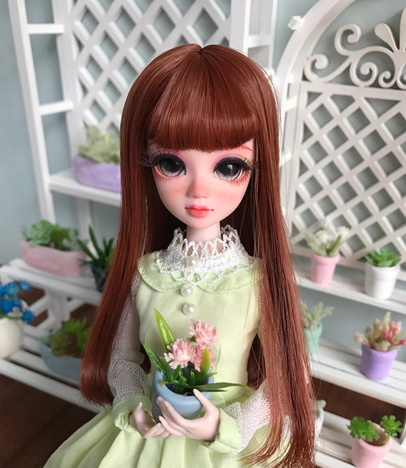 Puede incluir: Una mu&ntilde;eca con el pelo largo, liso y casta&ntilde;o, que lleva un vestido verde claro con un cuello de encaje blanco y sostiene una maceta peque&ntilde;a con flores rosas.