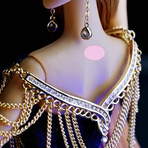 May include: A gold chain necklace with a diamond-encrusted pendant and a matching gold chain shoulder piece. The necklace is worn by a doll with long brown hair and a pink dress.
