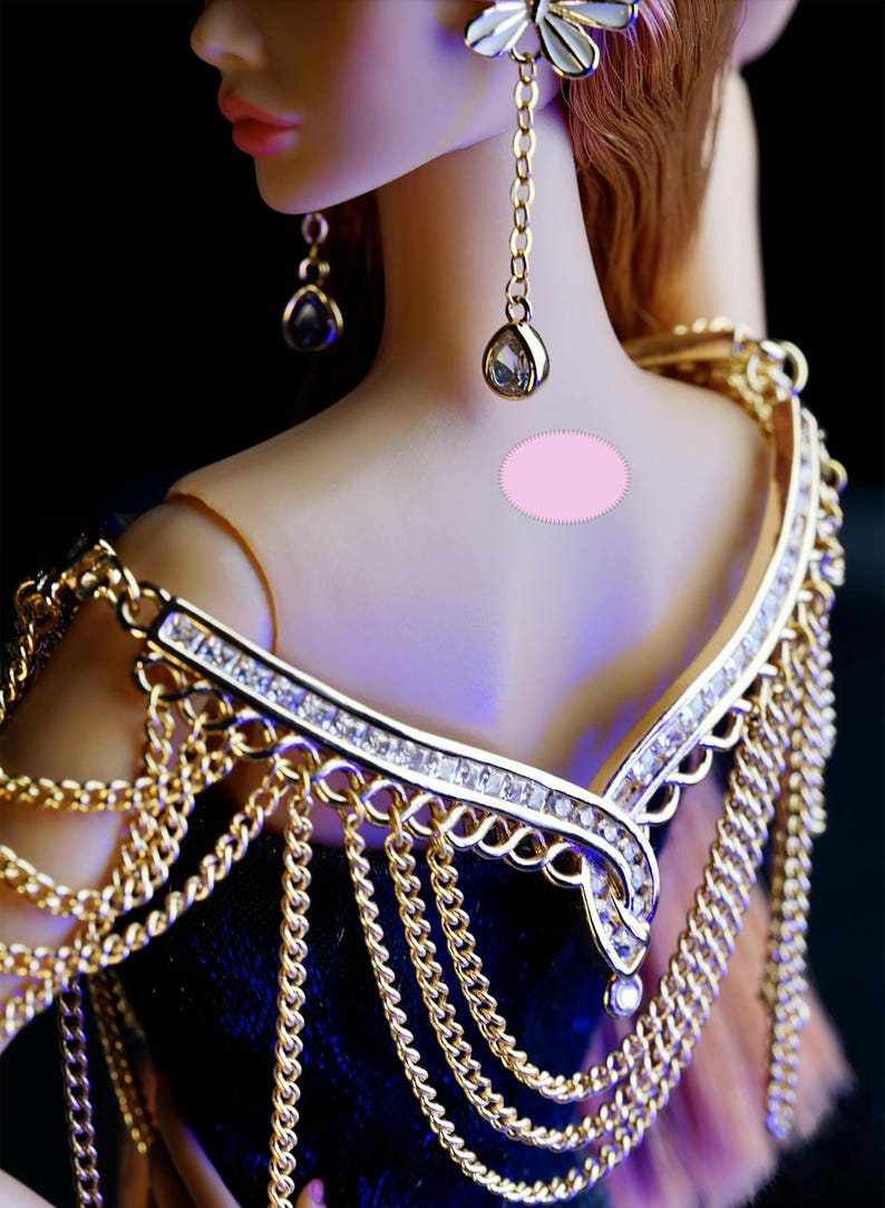 May include: A gold chain necklace with a diamond-encrusted pendant and a matching gold chain shoulder piece. The necklace is worn by a doll with long brown hair and a pink dress.