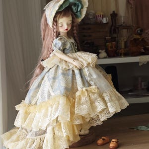 Puede incluir: Una mu&ntilde;eca de estilo vintage con pelo largo rosado, vestida con un vestido azul y crema con adornos de encaje. La mu&ntilde;eca tiene un gorro a juego con un lazo verde. Peque&ntilde;os zapatos marrones est&aacute;n colocados cerca.