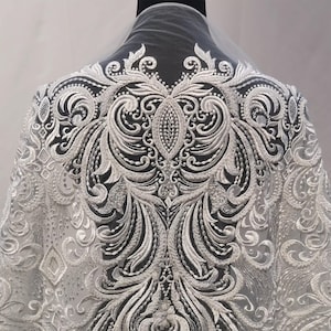 Puede incluir: Tela de encaje bordado blanco con un dise&ntilde;o ornamental y sim&eacute;trico. El patr&oacute;n presenta motivos florales y de volutas arremolinados, creando una apariencia detallada y texturizada. El encaje est&aacute; drapeado sobre un maniqu&iacute; negro.