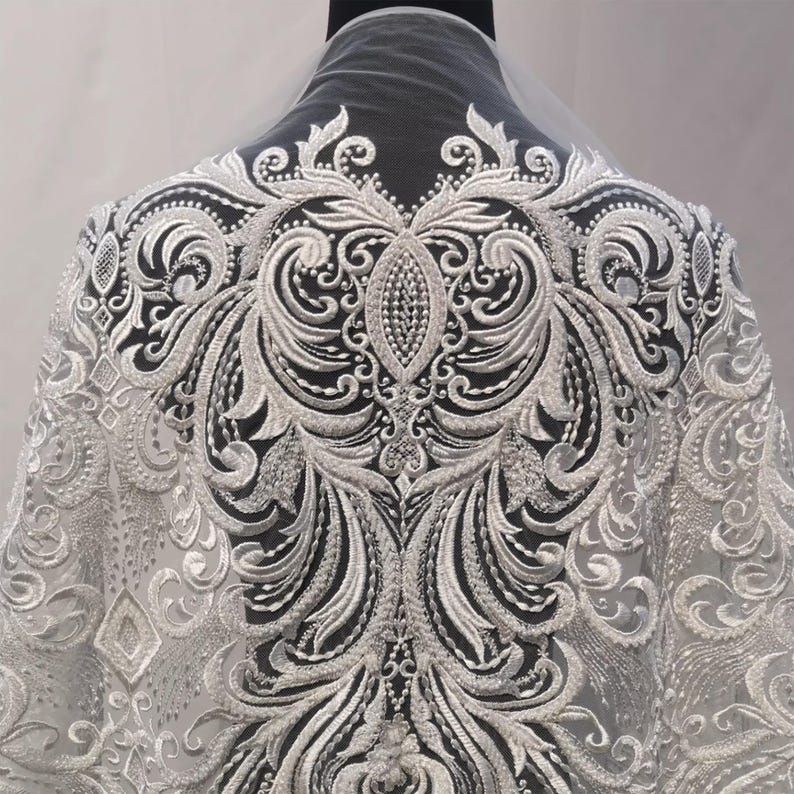 Puede incluir: Tela de encaje bordado blanco con un dise&ntilde;o ornamental y sim&eacute;trico. El patr&oacute;n presenta motivos florales y de volutas arremolinados, creando una apariencia detallada y texturizada. El encaje est&aacute; drapeado sobre un maniqu&iacute; negro.