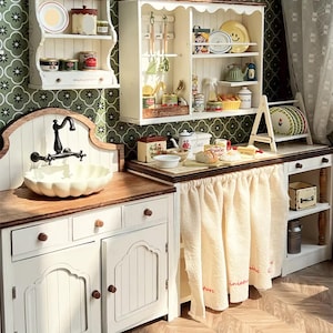 Puede incluir: Una escena de cocina en miniatura con armarios blancos, encimeras de madera y estanterías decorativas. La escena incluye un fregadero, un grifo y varios accesorios de cocina como platos, utensilios y tarros. El fondo presenta un papel pintado floral verde.