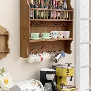 May include: A miniature kitchen scene with a wooden shelf holding bottles, cups, and mugs. A yellow espresso machine, a black coffee maker, and a white bread bin with floral details are visible. The bread bin has the text "Life is good!"