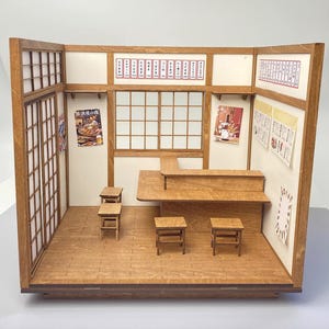 May include: A miniature wooden Japanese restaurant diorama with a wooden floor, walls, and furniture. The interior features small tables, a counter, and decorative posters. The walls have a white and beige colour scheme.