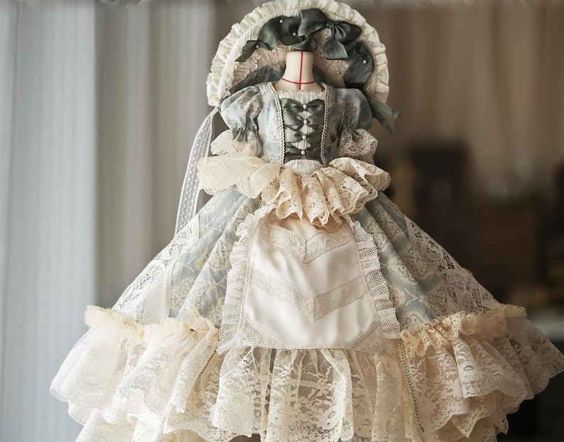 Puede incluir: Vestido de estilo vintage con un gorro, exhibido en un maniqu&iacute;. El vestido presenta un estampado floral azul claro y blanco, ribetes de encaje y una falda con volantes. El gorro est&aacute; adornado con lazos y encaje, creando una est&eacute;tica rom&aacute;ntica.