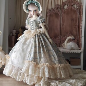 Puede incluir: Una mu&ntilde;eca de porcelana con un vestido de estilo vintage. El vestido presenta un corpi&ntilde;o con estampado azul y blanco, un ribete de encaje color crema y una falda larga y fluida. La mu&ntilde;eca lleva un gorro a juego con una cinta azul. La mu&ntilde;eca est&aacute; sobre una superficie de madera.