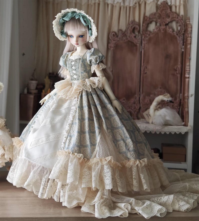 Puede incluir: Una mu&ntilde;eca de porcelana con un vestido de estilo vintage. El vestido presenta un corpi&ntilde;o con estampado azul y blanco, un ribete de encaje color crema y una falda larga y fluida. La mu&ntilde;eca lleva un gorro a juego con una cinta azul. La mu&ntilde;eca est&aacute; sobre una superficie de madera.