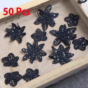 May include: A collection of black lace flower appliques with sequin detailing, displayed on a wooden tray. The text "50 Pcs" is visible. These appliques are suitable for various craft projects and embellishments.