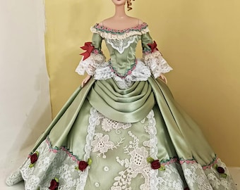 May include: A porcelain doll wearing a green satin gown with white lace trim and red rose accents. The gown has a full skirt and a fitted bodice. The doll has a blonde updo and a white face.
