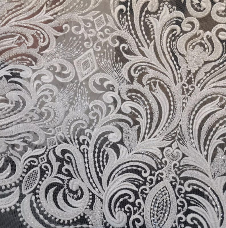 Puede incluir: Primer plano de tela de encaje blanco con un intrincado dise&ntilde;o floral y de volutas. El delicado patr&oacute;n presenta bordados detallados y acentos brillantes, creando una textura lujosa y elegante. El encaje se encuentra sobre un fondo oscuro.