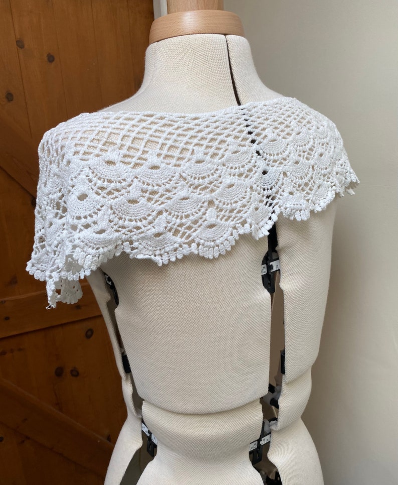 May include: White crochet lace collar with a scalloped edge. The collar is displayed on a white dress form.