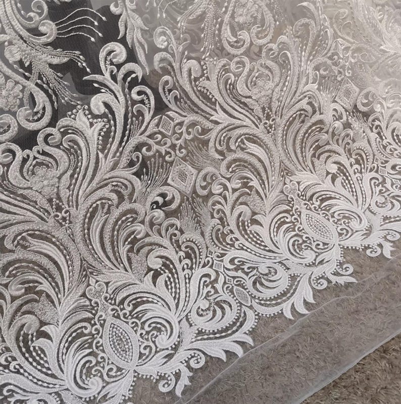 Puede incluir: Primer plano de tela de encaje blanco con un intrincado dise&ntilde;o floral y de volutas. El delicado bordado crea una superficie texturizada, perfecta para ropa de novia o manualidades elegantes. El patr&oacute;n es detallado y sim&eacute;trico.