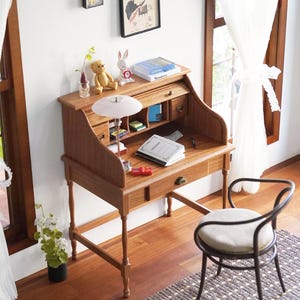 May include: A miniature wooden roll-top desk with a small chair. The desk features drawers, a pull-out writing surface, and is decorated with a lamp, books, and ornaments. The chair has a metal frame and a round, cushioned seat.