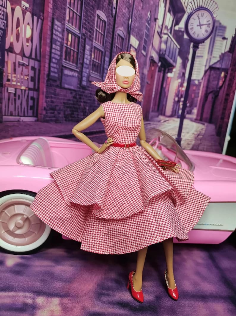K&ouml;nnte beinhalten: Eine Puppe in einem rot-wei&szlig; karierten Kleid mit rotem G&uuml;rtel und rotem Kopftuch. Die Puppe steht neben einem rosafarbenen Auto.