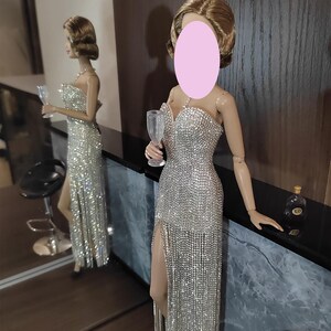 May include: A doll in a dazzling silver evening gown with a high split and a champagne glass. The dress is adorned with shimmering details. The doll is positioned near a mirror, reflecting the gown and the surroundings.